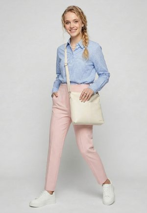 Junge Frau in hellblau kariertem Hemd, pinken Hosen, weißen Sneakers und cremefarbener Umhängetasche, lächelnd mit einer Hand in der Tasche.