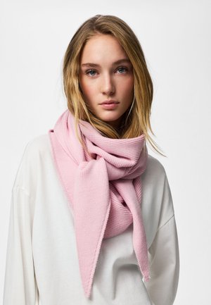 Tânără cu păr blond, purtând un fular mare tricotat roz și un top alb lejer, privind înainte pe un fundal simplu și luminos.