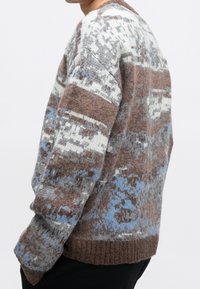 Personne portant un pull à motifs duveteux dans des tons marron, gris, blanc et bleu, vue de profil et de la taille vers le haut sur un fond uni.