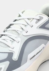 Jasnoszare buty sportowe z teksturowanej siateczki, białe akcenty oraz wyraźne logo "UNDER ARMOUR" na bocznej panelu.