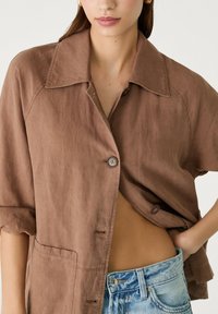 Kvinna iklädd en brun skjorta med knappar och uppvikta ärmar samt ljusblå denimjeans, med handen vilande på höften.