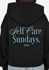 Čierna mikina s kapucňou so svetlomodrým textom na zadnej strane, ktorý znie "Self Care Sundays. Pegador." Tkanina vyzerá mäkko a má voľný strih.