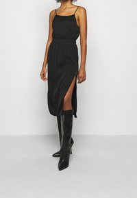 Robe midi en satin noir avec des bretelles fines, taille cintrée et fente sur le côté. Associée à des bottes noires à talon aiguilles jusqu'aux genoux. Texture lisse dans l'ensemble.