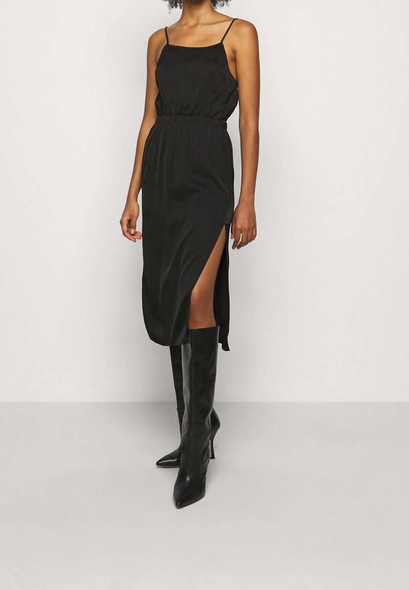 Robe midi en satin noir avec des bretelles fines, taille cintrée et fente sur le côté. Associée à des bottes noires à talon aiguilles jusqu'aux genoux. Texture lisse dans l'ensemble.