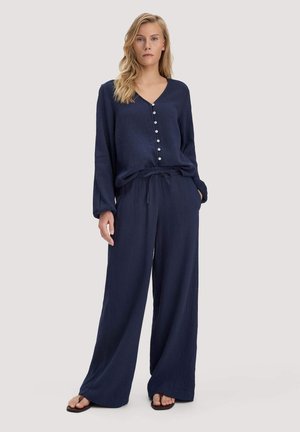 Frau trägt lockeres, marineblaues, langärmeliges, geknöpftes Oberteil und passende weit geschnittene Hose, steht mit einer Hand in der Tasche vor hellem Hintergrund.