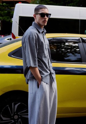 Mladý muž v slunečních brýlích a kostkované košili stojí s rukama v kapsách vedle žlutého auta na ulici.