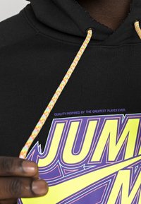 Felpa nera con cappuccio, dotata di cordini testurizzati multicolori. Presenta una grande grafica viola e gialla con la scritta "JUMP" e il testo sopra sul davanti.