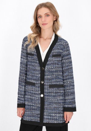 Tweed cardigan sinisenä ja valkoisena mustilla yksityiskohdilla, jossa on taitettu kaulus, etupainikkeet ja kaksi tikkipainetta.