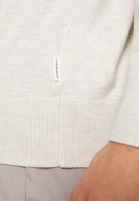 Ljus beige stickad tröja med en texturerad väv, ribbad fåll och liten märkesetikett på sidan. Mjuk och figurnära design.