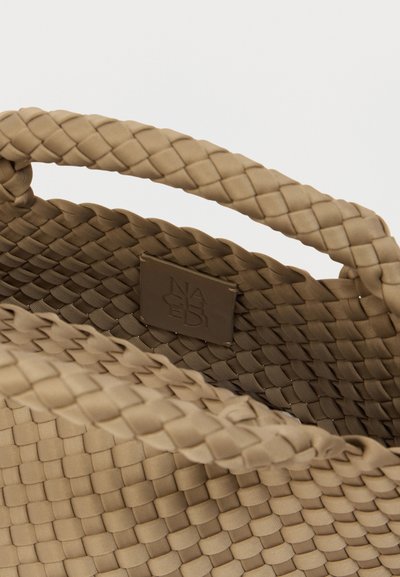 Πλεκτή μπεζ τσάντα με υφή, με στιβαρές λαβές και ετικέτα από δέρμα με εμπορικό σήμα στο εσωτερικό. Το υλικό φαίνεται συνθετικό και ευέλικτο.