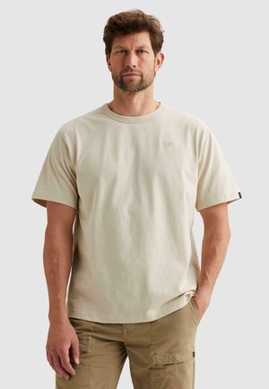 Man met kort bruin haar en baard, gekleed in een beige T-shirt en kaki cargobroek, staand met één hand in zijn zak.