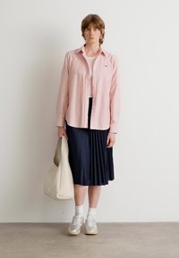 Camicia rosa chiaro a maniche lunghe, gonna a pieghe blu navy, sneakers grigie e beige, calzini bianchi e borsa a tracolla beige, in piedi su un pavimento neutro.