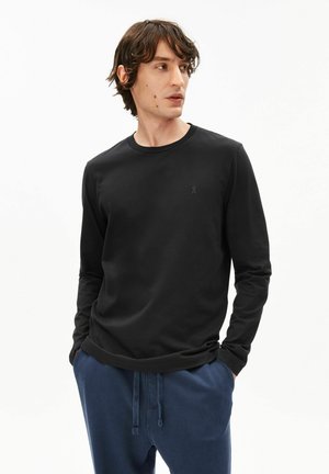 Mann trägt ein einfaches schwarzes langärmeliges Hemd und blaue Hosen, steht mit den Händen in den Taschen und schaut nach links vor einem weißen Hintergrund.