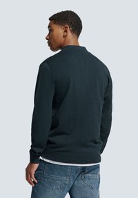 Donkergroen gebreid sweater met een ronde hals, geribbelde boorden en zoom, gecombineerd met lichtblauwe jeans. De stof lijkt zacht en gestructureerd.