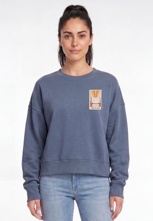Femme aux cheveux foncés en queue de cheval haute portant un sweat bleu avec un petit patch coloré d'un livre ouvert et le texte "The Reader" sur la poitrine.