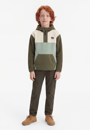 Enfant roux debout, portant un sweat à capuche polaire bicolore vert et beige, un pantalon cargo olive et des bottes à lacets couleur beige, sur fond blanc.