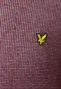 Tejido texturizado color burdeos con un patrón tejido; presenta un logo de águila bordado en amarillo, contorneado en negro.