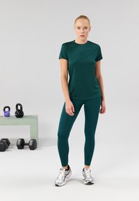 Teal athletic t-shirt and leggings in a smooth, stretch fabric; model standing with kettlebells and dumbbells in the background.