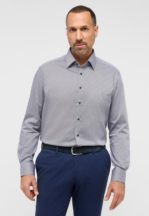 Hemd in Button-Up-Style mit einem blau-weißen geometrischen Muster, langen Ärmeln und einem spitz zulaufenden Kragen. Kombiniert mit navy blauen, strukturierten Hosen und einem schwarzen Gürtel.