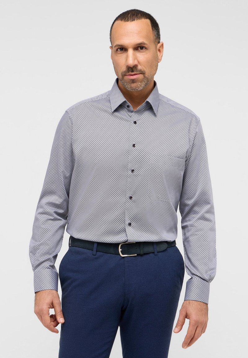 Overhemd met knopen en een blauw-witte geometrische print, lange mouwen en een puntige kraag. Gecombineerd met marineblauwe textuurbroek en een zwarte riem.