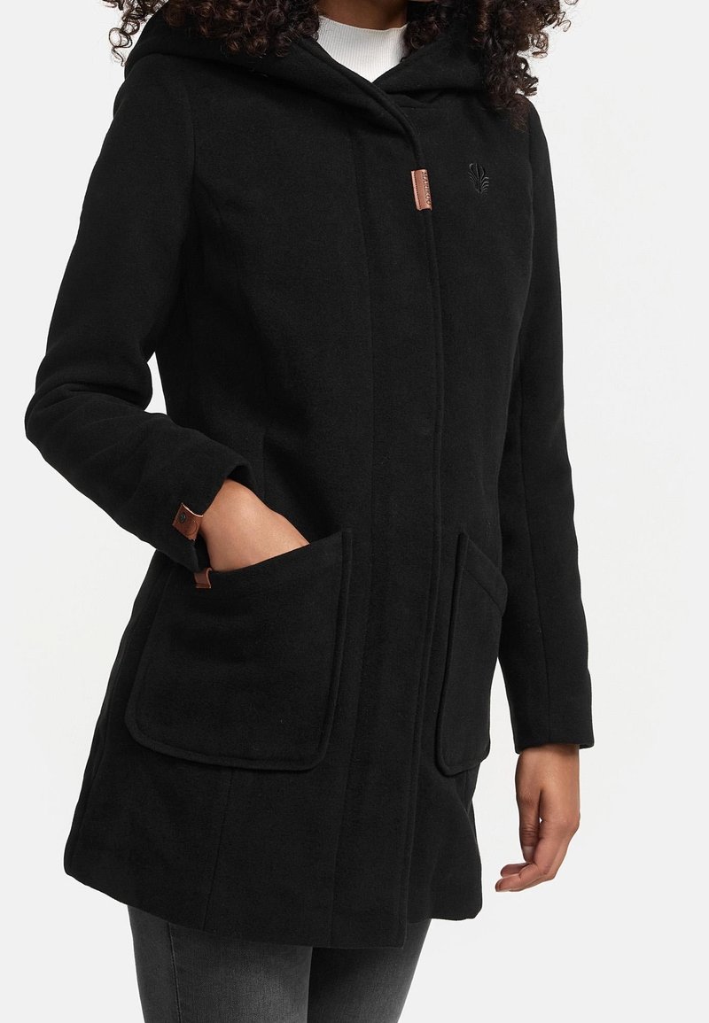 Femme portant un manteau noir à capuche avec de grandes poches avant et des détails de boutons marron, debout avec une main dans la poche.