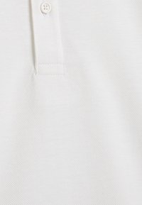 Camisa polo blanca con una superficie texturizada, que presenta un cuello con tapeta de dos botones y un sutil ribete diagonal en la tela.