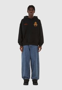 Felpa nera con una grafica sul petto, abbinata a jeans di denim blu oversize e sneakers nere.