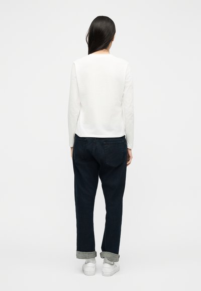 Γυναίκα που φαίνεται από πίσω, φοράει λευκό πουκάμισο με μακριά μανίκια, σκούρο τζιν με γυρισμένα μανίκια, γκρι κάλτσες και λευκά αθλητικά παπούτσια σε απλό φόντο.