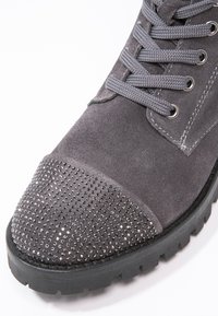 Botín de piel ante gris con punta redondeada, que presenta una puntera metálica con tachuelas, cordones grises y una suela de goma negra con patrones de tracción.
