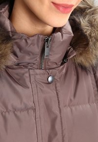 Brun puffjacka med en foderad huva, dragkedjestängning och tryckknappsstängning. Har en slät yta och quiltat mönster.