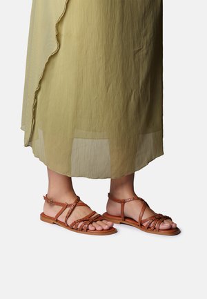 Nohy obuté v hnědých sandálech s pásky a plochou podrážkou pod průsvitnými, světle zelenými šaty ke kotníkům na jednobarevném pozadí.