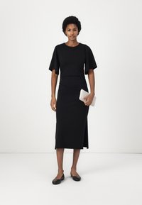 Black fitted dress with short flutter sleeves and a side slit. Model holds a textured light gray clutch, wearing black flat shoes.