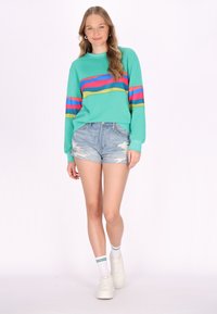 Mintgrüner Pullover mit mehrfarbigen horizontalen Streifen, kombiniert mit zerrissenen Denim-Shorts und weißen Turnschuhen, dazu Knöchelsocken.