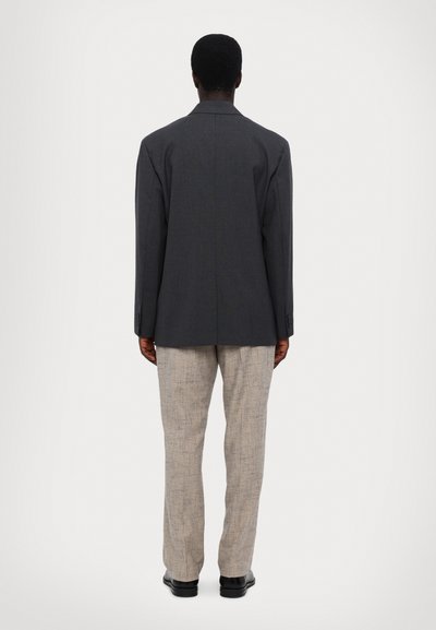 Ülevaatlik hall bleiser, millel on sile tekstuur, kaks tagumist lõhet, koos heleda beeži mustriga pükste ja mustade kingadega.