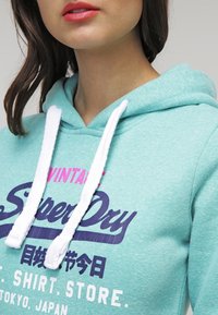 Felpa in cotone turchese con laccetti bianchi. Presenta il logo "SuperDry" in testo viola e rosa, insieme a caratteri giapponesi, sul fronte.