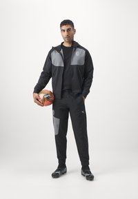 Zwarte sportieve trainingsoutfit met grijze accenten, bestaande uit een jasje met rits en broek met elastische taille. Model houdt een voetbal vast. Zwarte voetbalschoenen.
