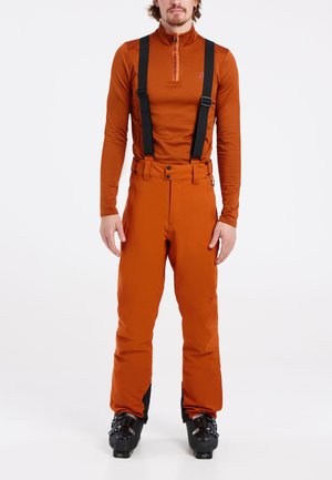 Pantalons de ski orange avec bretelles noires réglables, taille ajustée et une coupe légèrement détendue au niveau des jambes. Portés avec un haut à manches longues assorti.