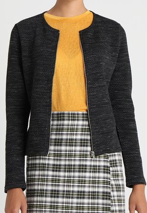 Veste en tricot noire avec un col rond, fermeture éclair à l'avant. Associée à un pull jaune et à une jupe à carreaux noire, verte et blanche.