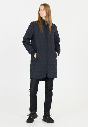 Nainen seisoo taskut käsissään pukeutuneena laivastonsiniseen keskipitkään tikattuun takkiin, mustiin farkkuihin ja mustiin vaelluskengissä yksiväristä taustaa vasten.