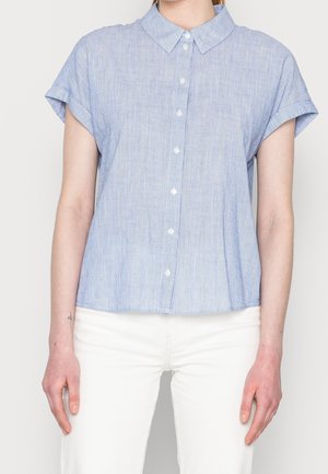 Mujer con camisa de botón de manga corta con finas rayas azules y blancas y pantalones blancos, de pie frente a un fondo liso.