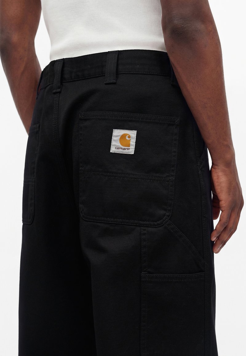 Person wearing black Carhartt pants with a white logo patch on the back pocket and a white sleeveless shirt.