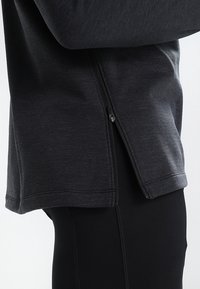 Haut de sport noir avec une petite fermeture éclair, en tissu texturé, ourlet arrondi et manches longues, associé à un pantalon noir moulant.