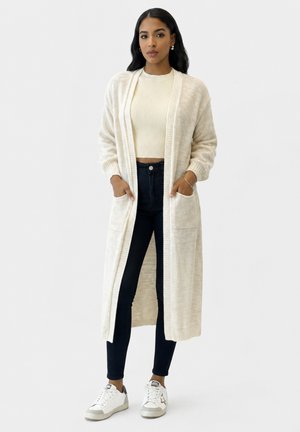Kvinde iført en lang cremefarvet cardigan, cremefarvet ribbet crop top, sorte skinny jeans og hvide sneakers med stjerne-detalje.