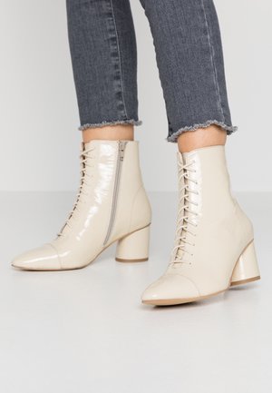 Beige patenterede læderankelstøvler med spidse tæer, snørebånd foran og lynlås i siden. Chunky hæl og en tekstureret denim-manchet er synlig.