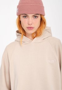 Beige hoodie met een trekkoordkap, zachte textuur en een klein geborduurd logo. Gecombineerd met een roze geribbelde beanie.