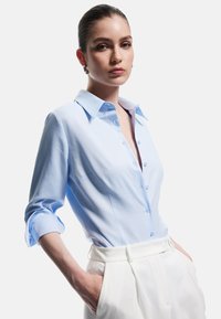 Light blue button-up shirt with a structured collar, rolled sleeves, and a smooth fabric finish, paired with high-waisted white trousers.
