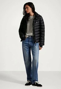 Γυναίκα με μαύρο μπουφάν, γκρι φούτερ, μπλε τζιν και μαύρα αθλητικά παπούτσια, που στέκεται με το ένα χέρι στην τσέπη και κοιτάζει στο πλάι.