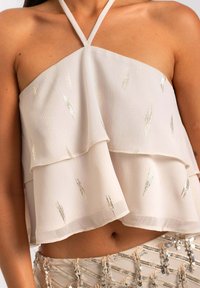 Femme portant un crop top à col halter blanc avec de subtils accents dorés et une jupe beige à sequins.