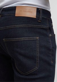 Gros plan de la poche arrière d’un jean en denim bleu foncé avec un patch en cuir marron portant la marque « 2Y PREMIUM » et des coutures orange visibles.