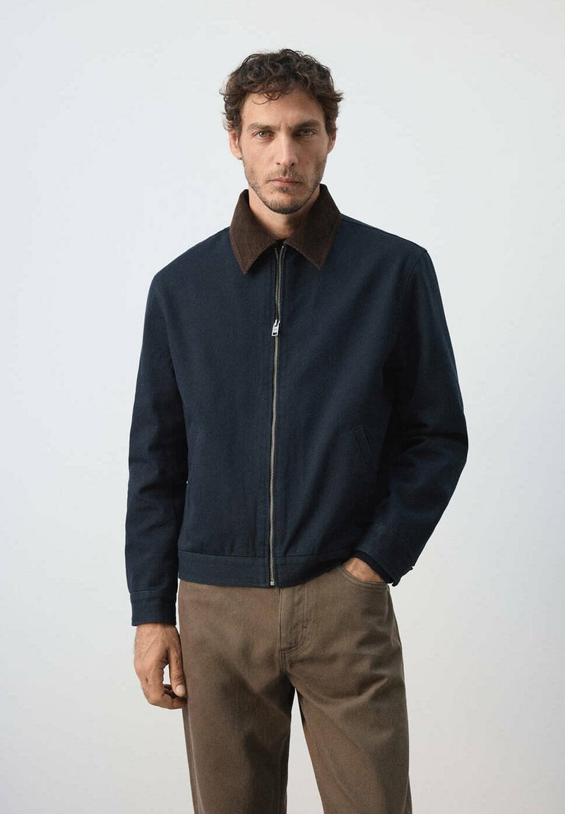Homme portant une veste bleu marine zippée avec un col marron et un pantalon marron, debout avec une main dans la poche devant un fond uni.